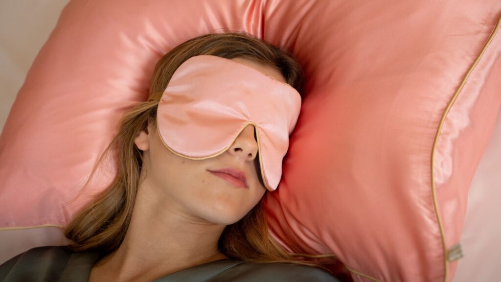 A person resting on a pink silk pillow while wearing a matching pink silk sleep mask