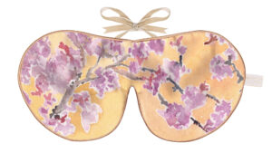 The image shows a silk sleep mask decorated with a botanical illustration inspired by hydrangea flowers. The front surface displays clusters of rounded hydrangea blossoms positioned across the mask in soft shades of pink, lavender, lilac, and pale cream. Each cluster is composed of many small petal shapes, rendered with layered watercolour-style shading and fine ink outlines that give a hand‑drawn, detailed effect. Between the hydrangea blooms are large, elongated leaves in varied shades of green, with textured vein lines that add depth. The background is a bright, clear blue wash, creating a vivid contrast against the softer floral colours and giving the design a fresh, summery quality. The mask is finished with warm-toned piping along the edge, outlining the curved silhouette. At the top, a beige ribbon is tied into a neat bow, indicating a ribbon‑tie fastening. A small fabric tag is attached at the right edge. The overall look is vibrant, botanical, and painterly, with an emphasis on floral detail and colour harmony.