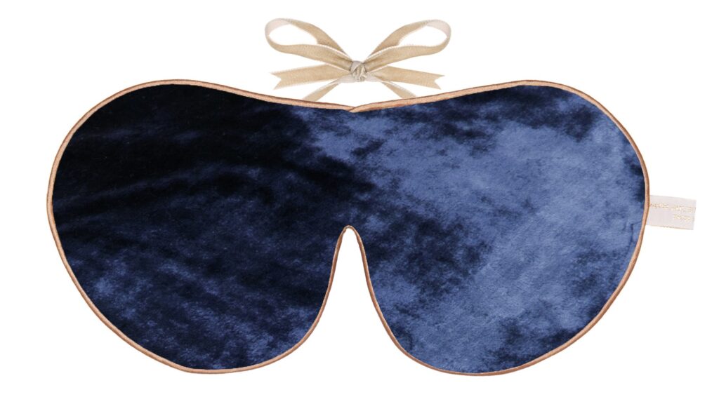 A navy blue silk velvet sleep mask with gold piping and soft beige ribbon ties.