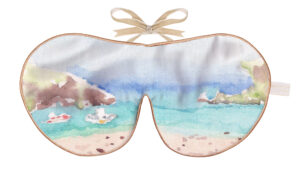 Silk sleep mask with a coastal scene showing turquoise water, sandy shoreline, and distant cliffs in soft watercolour tones.