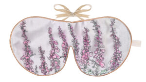 Silk sleep mask featuring a botanical illustration of tall pink foxglove flowers on a light background, finished with warm piping and a beige ribbon tie.