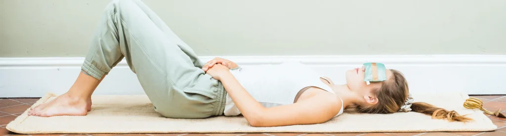 A person lying on their back on a fleece wellness rug mat, wearing a silk eye pillow, with knees bent and hands resting on their stomach.