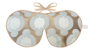 A silk sleep mask with a geometric woven pattern in soft blue and taupe tones, finished with gold edging and beige ribbon ties.