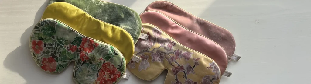 A selection of silk sleep masks arranged in a row, showing different colours and textures, laid out on a soft grey surface.