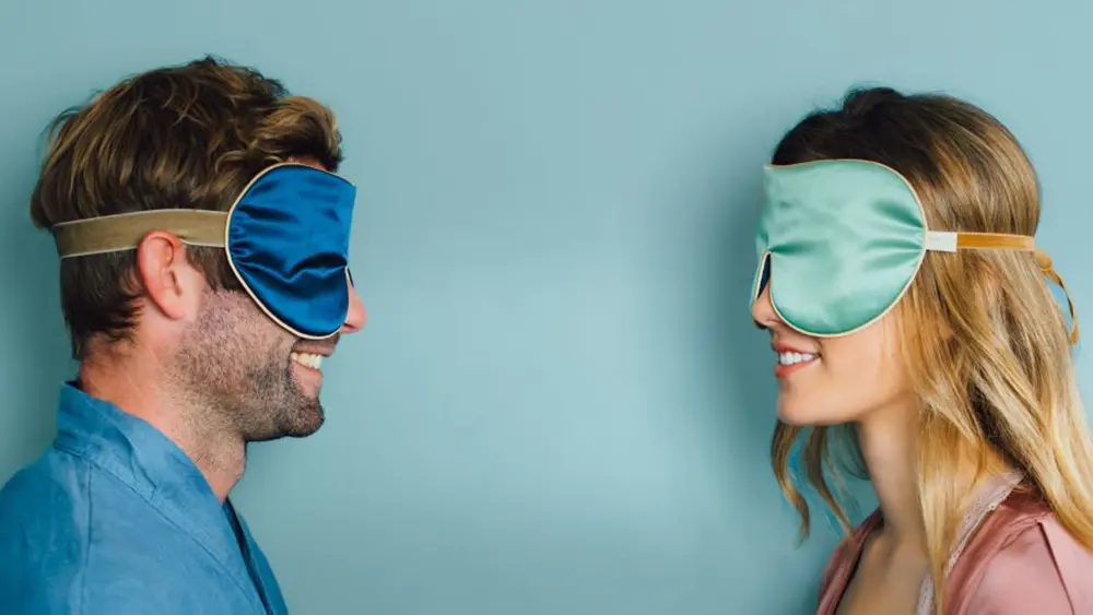 Two people facing each other against a soft blue background, each wearing a sleep mask 