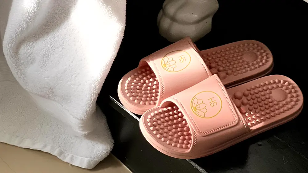 Pair of pink massage‑sole slide slippers with raised acupressure dots, placed on a black surface beside a white towel.